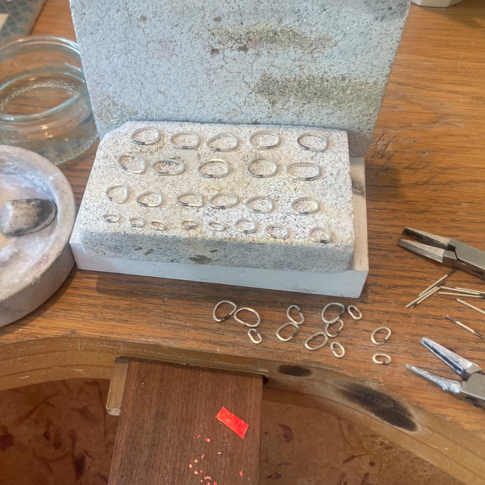 Jeweller’s bench with soldering blocks & numerous sterling silver square wire rings beside some pliers & solder pallions